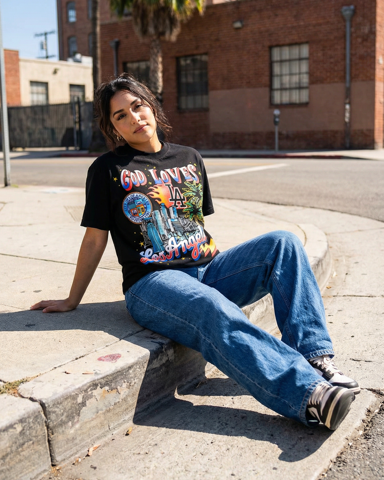 Person sitting on a street curb wearing a graphic t-shirt and jeans, with a brick building and palm tree in the background.